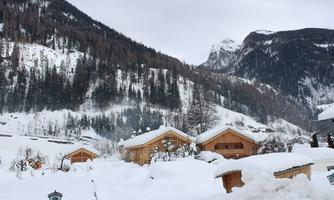 A winter landscape with snow-covered huts and majestic mountains in the background. The sky is gray, which enhances the serene atmosphere.