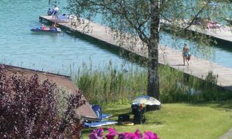 A tranquil lake with a dock and green meadows. Colorful flowers in the foreground and a few people by the water.