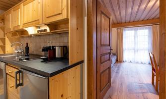 A cozy wooden kitchen with modern amenities. In the background, a bright room with large windows and a wooden floor can be seen.