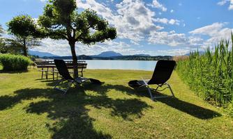 A quiet lake with deck chairs on a green meadow. In the background, mountains and a clear sky are visible.