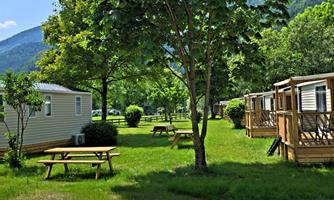 A quiet campsite with green meadows and large trees. Cozy mobile homes and picnic tables are nearby.