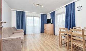 A modern living room with a beige sofa and a wooden dining table. Large windows provide plenty of daylight and blue curtains complete the room.