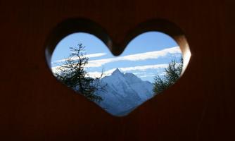 A view through a heart-shaped window at snow-covered mountains under a clear blue sky. The surroundings show some trees and a beautiful landscape.