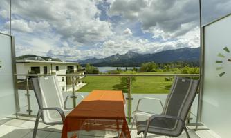 A balcony with two chairs and a table that overlooks a picturesque landscape with mountains and a lake. The sky is cloudy, giving the scene a tranquil atmosphere.