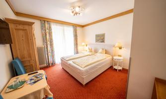 A cozy double room with shower/WC, large balcony, and colorful curtains. There is a table and a chair as well as a wooden wardrobe.