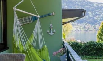 A quiet balcony with a hanging lounger and a beautiful view of a lake. In the background, green mountains and a suggested garden are visible.
