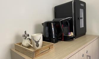 A cozy corner with a coffee machine, a kettle, and a cup on a wooden tray. Underneath is a container for milk.