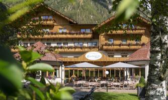 A cozy hotel with a wooden facade and balconies. The outdoor area is equipped with seating and umbrellas, surrounded by green trees.
