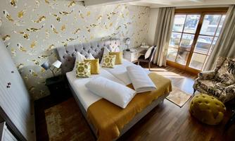 An elegant bedroom with a large bed, colorful pillows, and a cozy armchair. Large windows offer a view outside and let in plenty of daylight.