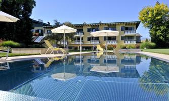 A clear pool with sun umbrellas and loungers in a well-kept garden. In the background, a modern building with balconies can be seen.