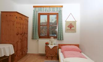 A cozy room with a single bed and a wooden wardrobe. Natural light comes in through the window, and there is a tablecloth on a small table.