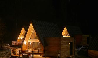 Three wooden chalets glow softly in the night. Surrounded by trees, they offer a cozy atmosphere.