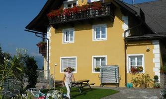 A charming yellow house with a balcony and flowers. In the foreground, you can see a person on the green lawn in front of the house.