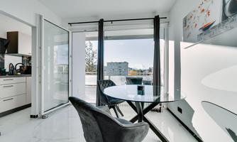 A modern dining room with a round glass table and a black chair. Large windows provide a lot of light and a view outside.