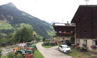 A rural landscape with several tractors and a red farmhouse. In the background, green mountains and wooded hills are visible.