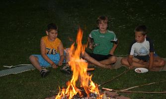 Three children are sitting by the campfire roasting marshmallows. The atmosphere is cozy and friendly.
