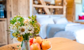 A beautiful table with a vase full of flowers and a bowl of apples. In the background, a cozy room with wooden details can be seen.