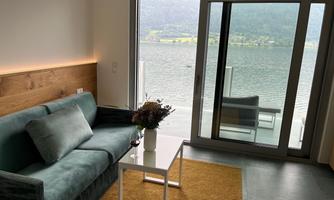 Modern living area with a green sofa and a small white table. Through the large windows, one looks out at a lake with a picturesque landscape in the background.