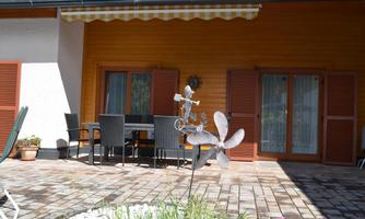 A cozy outdoor area with a table and chairs. In the foreground, a flower bed and a decorative wind chime.