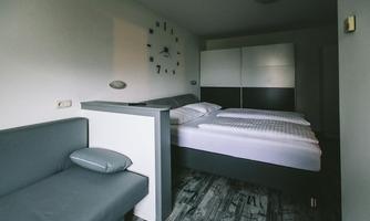 A modern bedroom with two beds and a gray couch. The walls are light, and a large clock hangs on the wall.