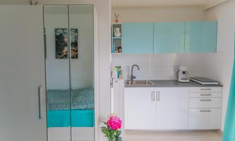 A modern kitchen unit with white furniture and turquoise accents. A cozy bed is visible in the background.