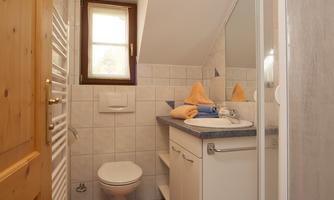 A modern bathroom with white tiles and a shower area. Orange towels and a mat give the room a friendly touch.