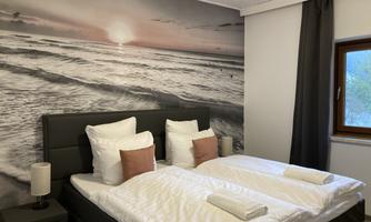 A cozy bedroom with two beds and a large wall mural of a sunset at the sea. The soft color scheme and the furnishings create a relaxed atmosphere.