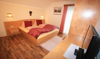 A cozy bedroom with a double bed and warm colors. Pictures hang on the wall, and a television is placed on a sideboard.