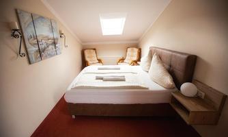 A cozy bedroom in the attic with a large bed and two chairs. The walls are light-colored and a window provides daylight.