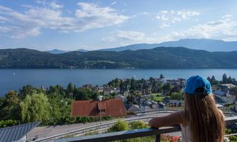 A girl with a blue cap is standing on a balcony and looking at a beautiful lake. In the background, there are mountains and a picturesque city.