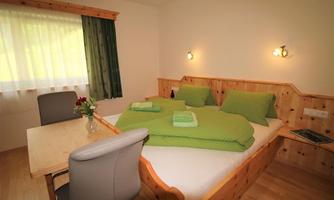 A cozy bedroom with a double bed and green bedding. Next to the bed is a small table with a flower vase and a chair.
