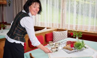 A smiling woman is setting a table with cups and a treat. In the background, there are windows with curtains and cozy seating areas visible.