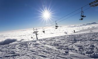 A wintry mountain landscape with lots of snow and ski lifts under a clear blue sky. The sun shines above the clouds.