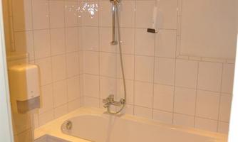 A simple bathroom with a white bathtub and a shower. The walls are tiled and the floor is in terracotta style.