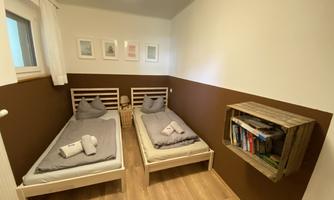 A cozy bedroom with two single beds and light walls. In one corner, there is a wooden shelf with books.