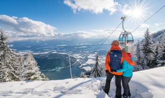A couple stands in the snow and enjoys the view of the mountains. In the background, you can see a cable car and a clear blue sky.
