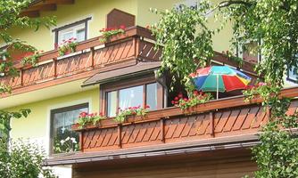A bright house with a balcony full of flowers. A colorful sun umbrella stands on the balcony under the trees.
