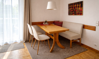 A cozy dining area with a wooden table and upholstered chairs. The light falls through large windows, creating an inviting space.