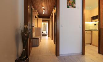 A bright hallway with wooden paneling and narrow doors. In the background, a kitchen is visible, and there are decorative elements like a painting and a plant vase.