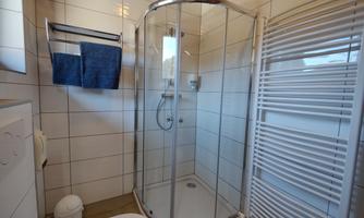 A modern bathroom with a shower cabin and white tiles. Towels are hanging on the wall and a towel warmer is visible.