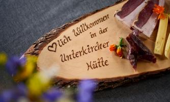 A wooden board with the inscription "Welcome to the Unterkircher Hut". On it are various types of sausage and cheese as well as fresh flowers.
