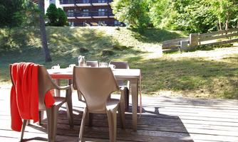 A cozy table with chairs on a wooden terrace. In the background, trees and a building can be seen.