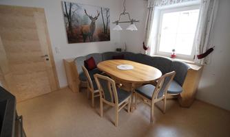A cozy dining area with a round wooden table and a U-shaped bench. A picture of a deer hangs on the wall and the window provides bright light.