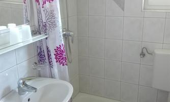 A small bathroom with a shower, sink, and toilet. The walls are decorated with white tiles, and the shower curtain has a floral pattern.