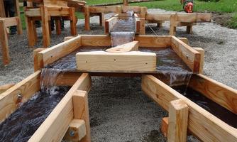 A water feature with wooden channels, the water flows through various sections. The environment is green and natural.