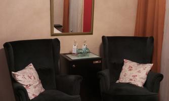 English
Two dark green wingback armchairs with cushions featuring a rose motif, a minibar with drinks between them; a wall mirror above, curtains on the right, and a bed with a rose pattern in the foreground.
