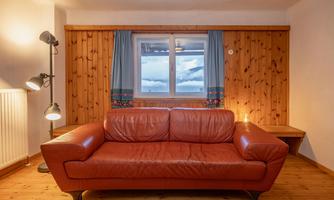 A cozy room with a brown leather sofa and wood paneling. There is a window with blue curtains that opens up the view outside.