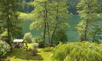 A peaceful landscape with a clear lake and lush greenery. In the foreground, trees and a small seating area are visible.