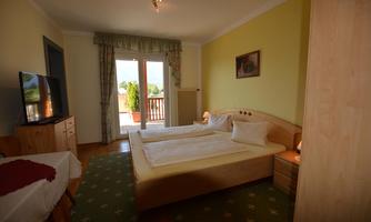 A bright bedroom with two beds, a television, and a balcony. The walls are painted yellow, and there is a carpet with a simple pattern.
