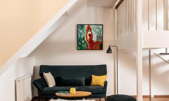 A modern living room with a dark green sofa and a round coffee table. A colorful painting hangs on the wall, and the floor is made of light wood.